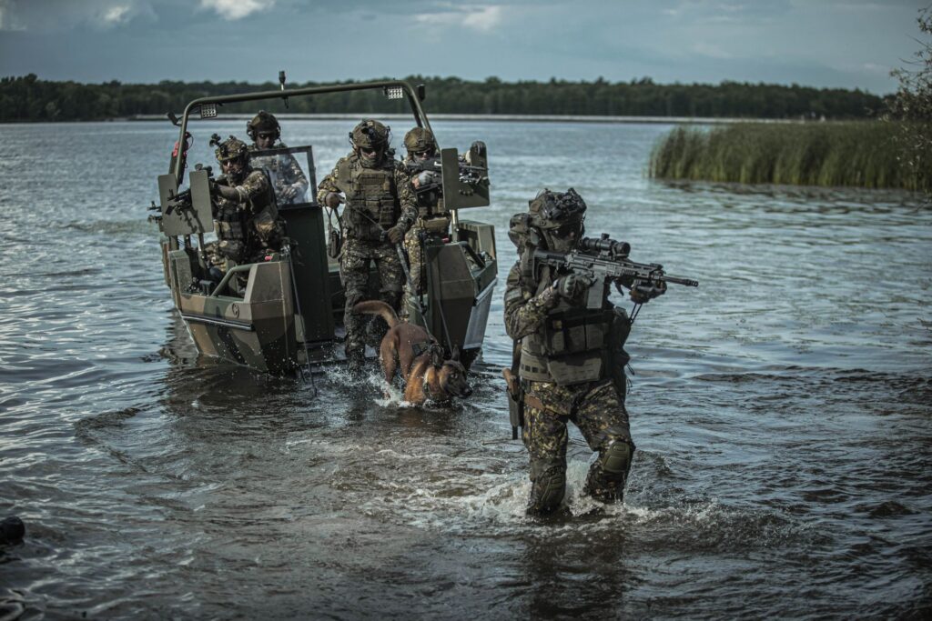 Łódz militartna na wodzie, z żołnierzami, z ciężką bronią i psem wychodzącym po rampie na brzeg.